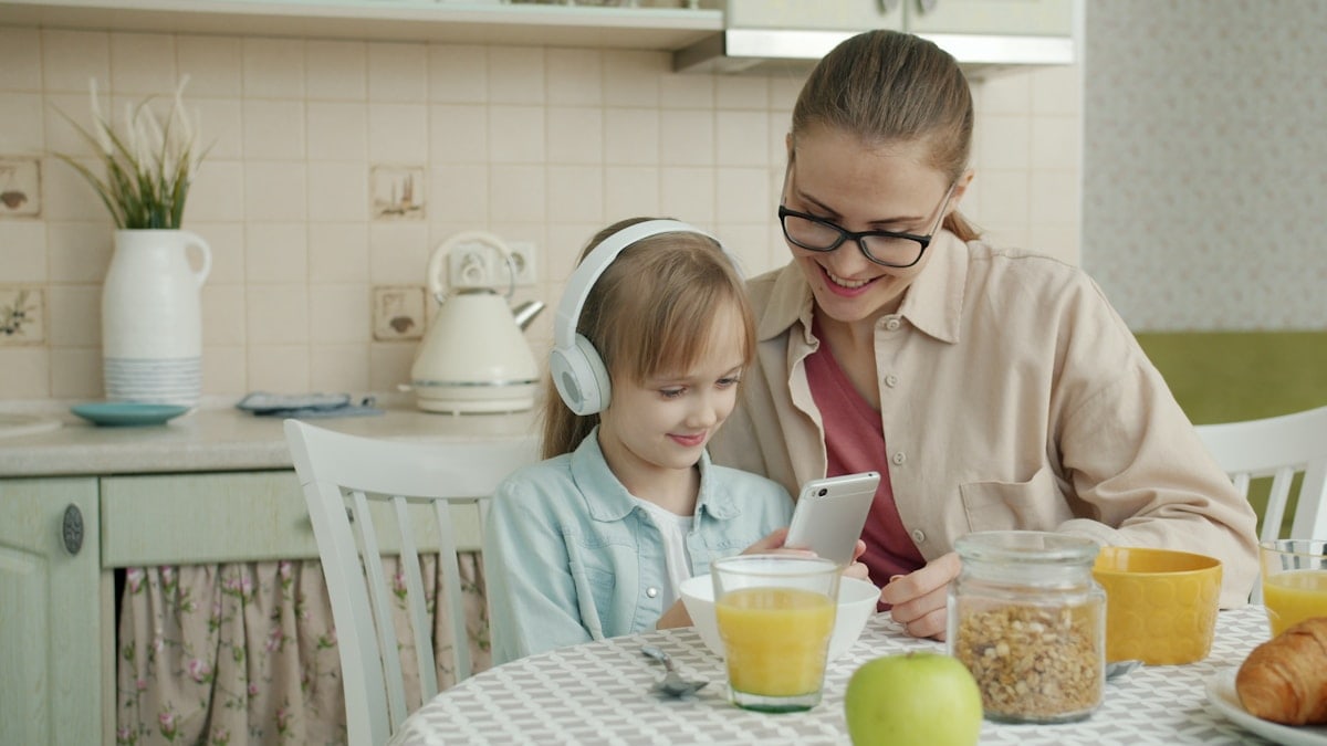 Slow motion of happy little girl and loving mother using smartphone and listening to music in headphones at kitchen table. Childhood and motherhood concept.